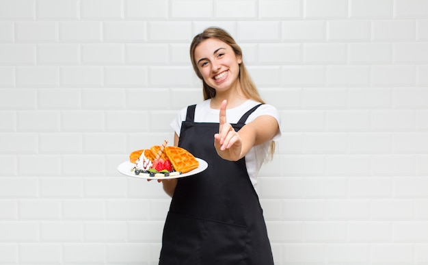 Mulher loira bonita sorrindo com orgulho e confiança fazendo a pose número um triunfantemente, sentindo-se uma líder