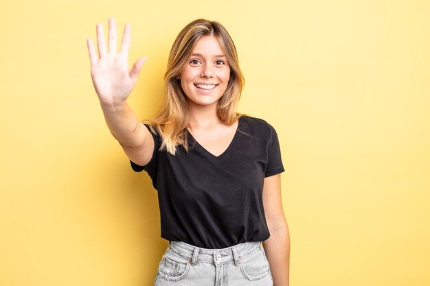 Mulher loira bonita sorrindo e parecendo amigável, mostrando o número cinco ou quinto com a mão para a frente, em contagem regressiva