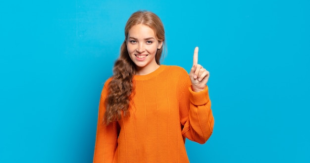 Mulher loira bonita sorrindo e parecendo amigável, mostrando o número um ou primeiro com a mão para a frente, em contagem regressiva