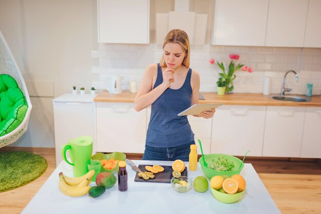 Mulher loira bonita vegan procurando uma receita no tablet para cozinhar frutas orgânicas na cozinha. Comida saudável. Desintoxicação da dieta