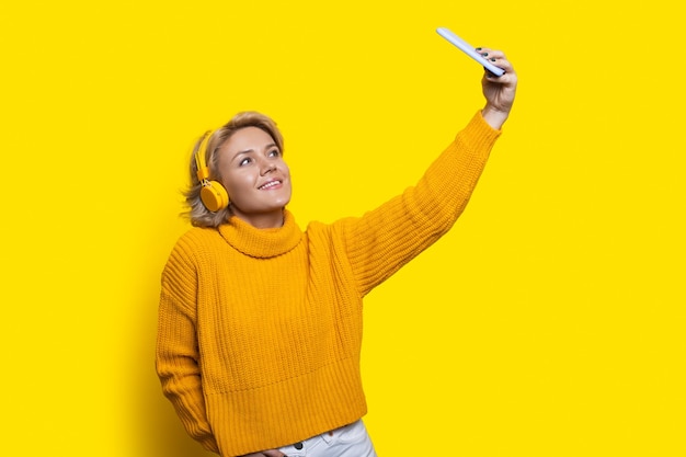 Mulher loira caucasiana está sorrindo em uma parede amarela fazendo uma selfie com o telefone e usando fones de ouvido