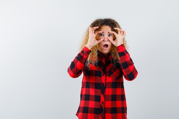 Mulher loira com cabelo encaracolado, abrindo os olhos com os dedos em uma camisa xadrez e parecendo chocada. vista frontal.