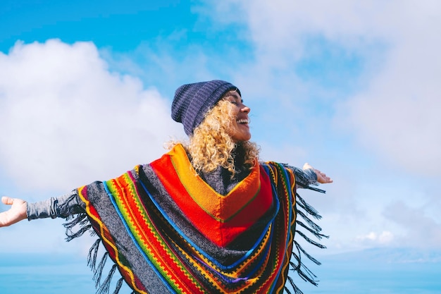 Mulher loira com chapéu e poncho colorido sorri e relaxa com os braços abertos no céu azul de inverno o estilo de liberdade do povo