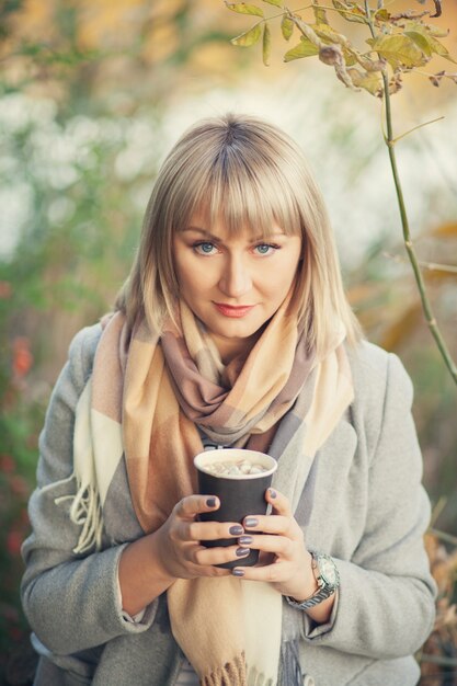 Mulher loira com corte de cabelo curto em um casaco de lã cinza e lenço xadrez bebe cappuccino de café com marshmallows. Caminhar e fazer um piquenique na natureza na floresta de outono