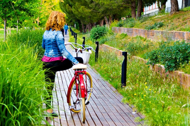 Mulher loira com jeans azul passeando no parque em uma bicicleta vermelha.