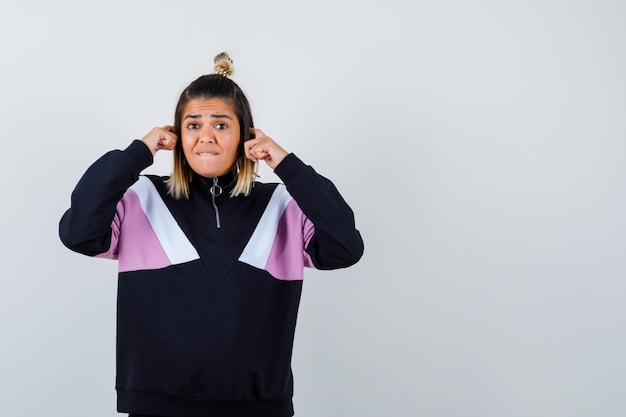 Mulher loira com um agasalho de treino preto segurando os dedos nas orelhas e parecendo confusa