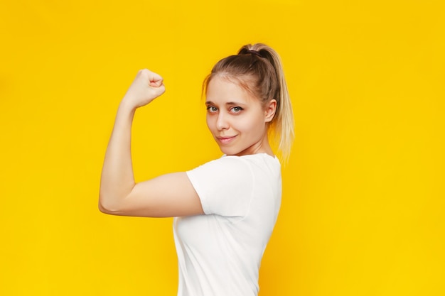 Mulher loira confiante, poderosa e poderosa, levanta o braço e mostra o bíceps isolado em uma parede amarela colorida brilhante