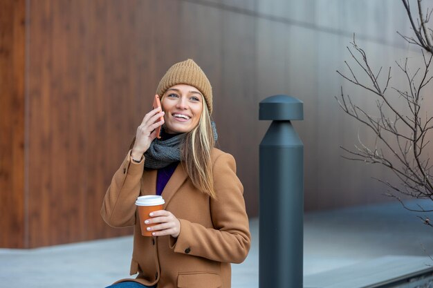 Mulher loira conversando navegando na internet on-line no smartphone durante a pausa para o café enquanto relaxa no banco do parque Linda jovem bonita usando telefone celular e segurando a xícara de café