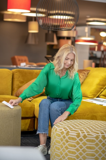 Mulher loira de blusa verde e calça jeans, sentada em um sofá amarelo