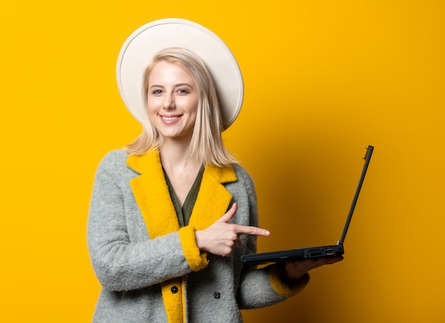 Mulher loira de chapéu e casaco com laptop na parede amarela