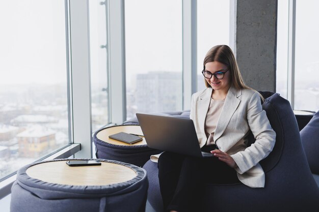 Mulher loira de conteúdo atraente em roupas casuais e óculos concentrando-se na tela e digitando no laptop enquanto está sentado no pufe macio na sala de estar