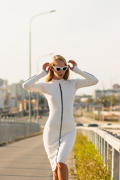 Mulher loira de corpo inteiro em elegante vestido branco olhando para longe enquanto está no caminho de asfalto