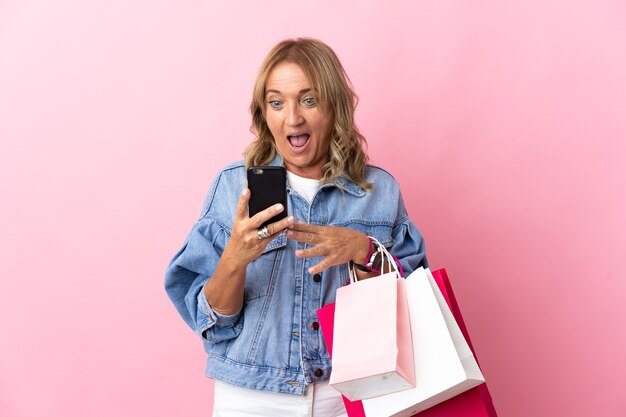 Mulher loira de meia-idade sobre um fundo rosa isolado segurando sacolas de compras e escrevendo uma mensagem com seu telefone celular para um amigo