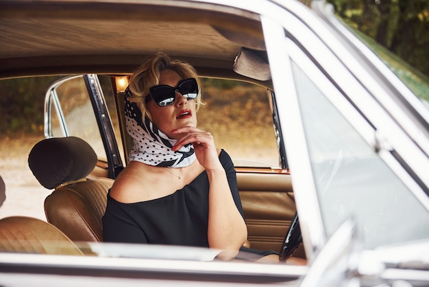 Mulher loira de óculos escuros e vestido preto, senta-se em um carro antigo clássico vintage.