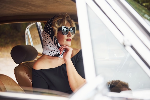 Mulher loira de óculos escuros e vestido preto, senta-se em um carro antigo clássico vintage.