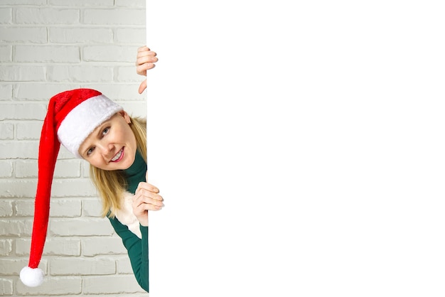 Mulher loira de Papai Noel de Natal espiando por trás do outdoor de sinal em branco atribuir a parede de tijolos brancos. Natal