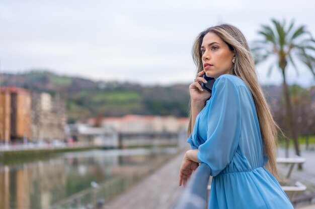 Mulher loira de vestido azul com telefone no conceito de estilo de vida do rio da cidade