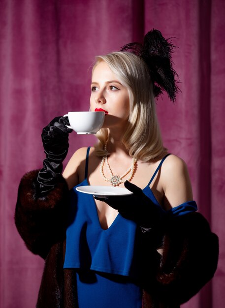 Mulher loira de vestido azul vintage e casaco de pele com uma xícara de café