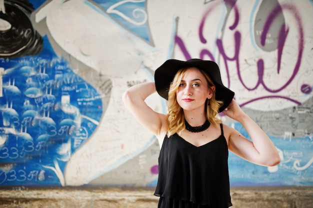 Mulher loira de vestido preto, colares e chapéu contra parede grafite.