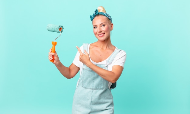 Mulher loira e bonita sorrindo alegremente, sentindo-se feliz e apontando para o lado e pintando uma parede