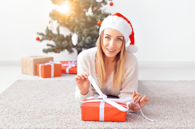 Mulher loira e bonita, vestindo um suéter de malha quente, deitada sobre um tapete perto da árvore de Natal decorada em uma sala de estar. Muitos presentes debaixo da árvore.