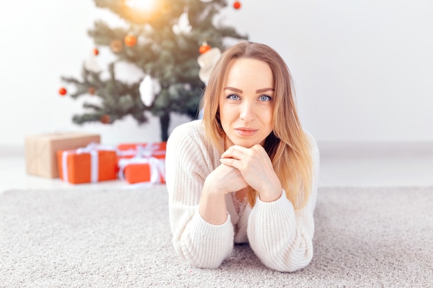 Mulher loira e bonita, vestindo um suéter de malha quente, deitada sobre um tapete perto da árvore de natal decorada em uma sala de estar. muitos presentes debaixo da árvore.