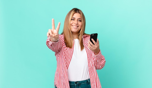Mulher loira e curvilínea sorrindo e parecendo amigável, mostrando o número dois e segurando um telefone inteligente