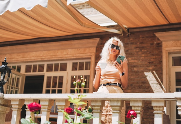 Foto mulher loira elegante adulta em óculos de sol e roupas bege usando telefone celular no quintal de casa no café de rua da cidade de verão