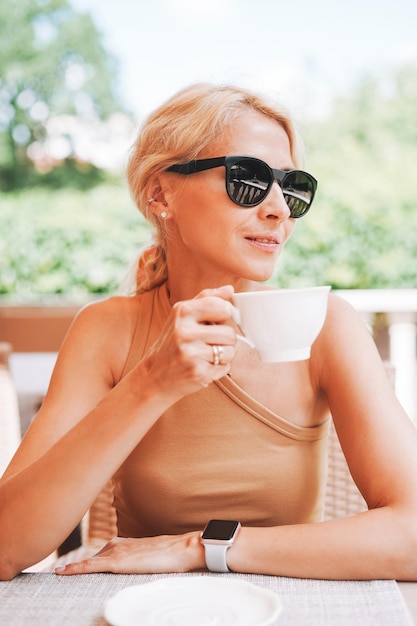 Mulher loira elegante adulta em óculos de sol roupas bege com uma xícara de café no café de rua da cidade de verão