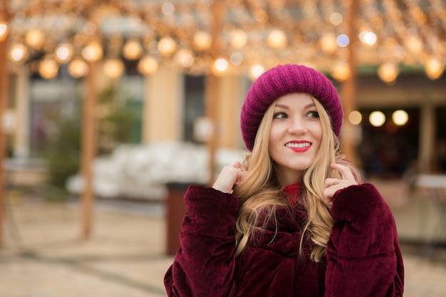 Mulher loira elegante com chapéu de malha vermelho e casaco de inverno, posando no fundo de uma guirlanda na rua em Kiev