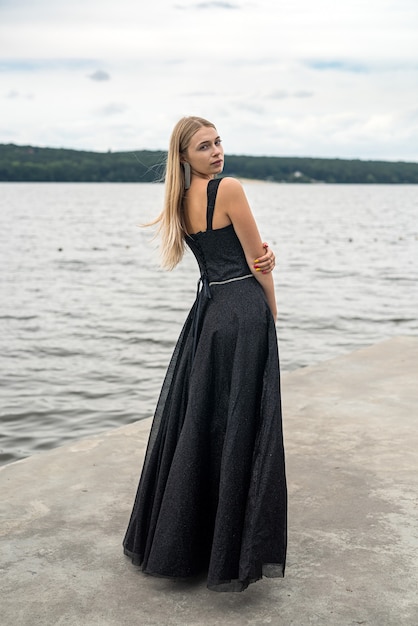 Mulher loira elegante em um vestido de noite preto da moda perto do lago, estilo de vida