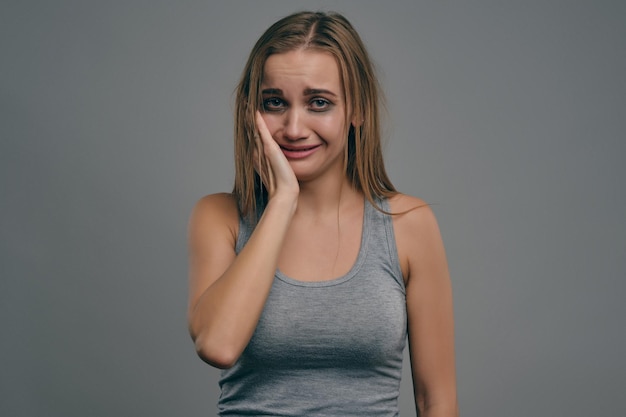 Mulher loira em pânico com cabelos despenteados e hematomas está tocando seu rosto, de camiseta cinza, posando em fundo cinza do estúdio. Violência doméstica, abuso. Depressão, desespero. Close-up, copie o espaço.