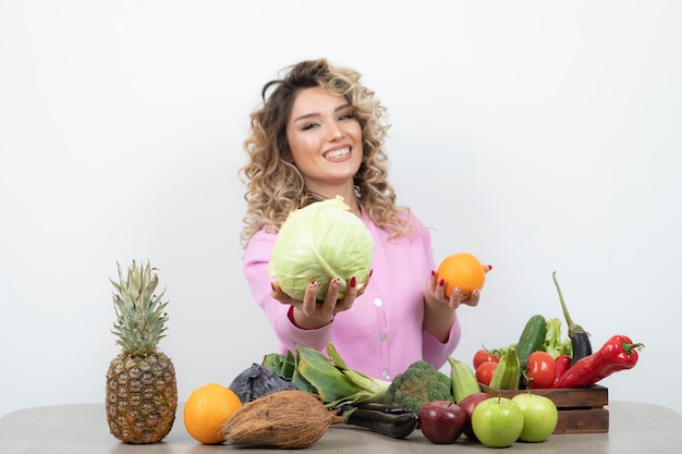 mulher loira em top rosa segurando repolho e laranja na mesa.