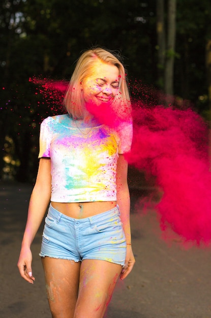 Mulher loira emocional em uma camiseta branca e shorts jeans brincando com o pó de Holi vermelho explodindo ao seu redor