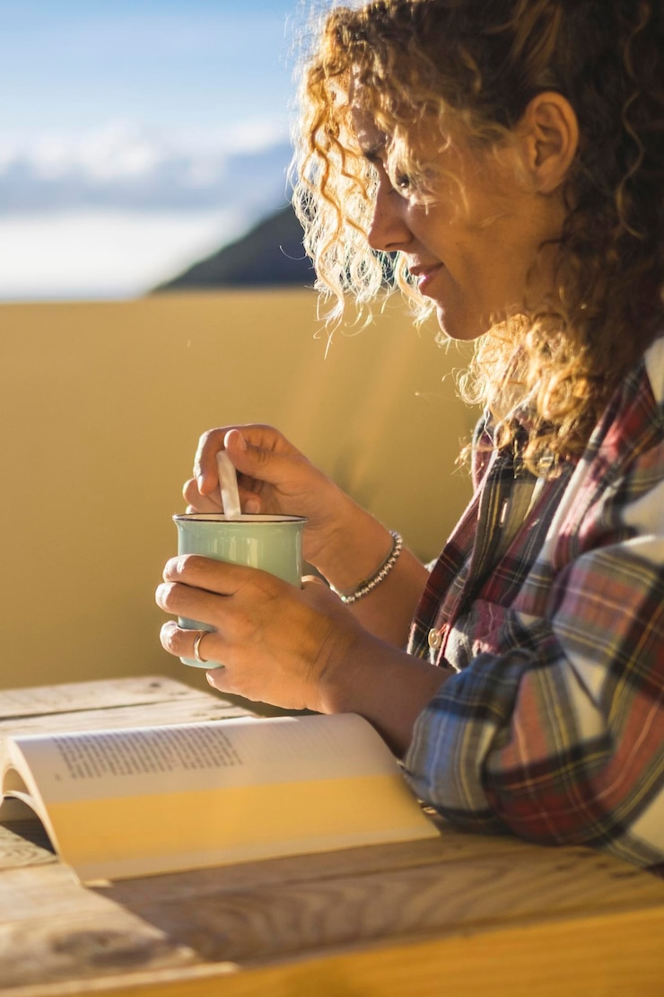 Foto mulher loira encaracolada relaxa lendo um livro sentada na mesa no terraço conceito de lazer e relaxamento