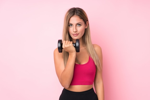 Mulher loira esporte jovem isolado rosa fazendo levantamento de peso com kettlebell e olhando para a frente