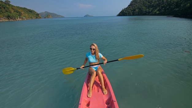 Mulher loira esportiva atraente com óculos de sol enfileira canoa de plástico rosa ao longo da água do mar