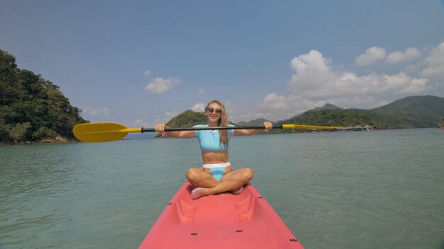 Mulher loira esportiva atraente com óculos de sol enfileira canoa de plástico rosa ao longo da água do mar