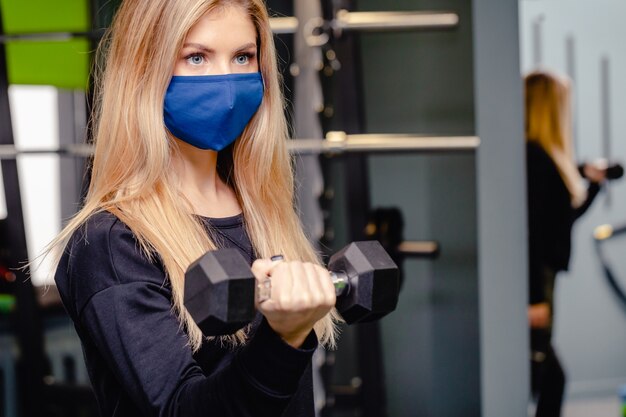 Mulher loira está treinando na academia durante a pandemia. menina faz exercícios com máscaras médicas. ginásio durante o período do coronavírus. coronavírus, doença, conceito de infecção.