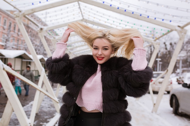 Mulher loira expressiva com casaco de pele quente posando com o vento no cabelo na cidade no inverno
