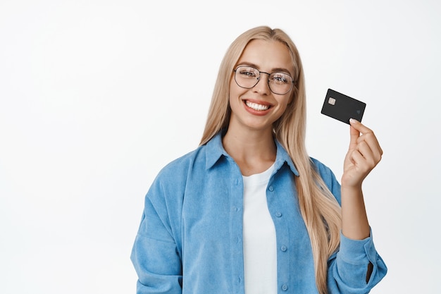 Mulher loira feliz de óculos sorrindo, mostrando o cartão de crédito na mão, recomendando se tornar cliente do membro do banco, em pé no branco