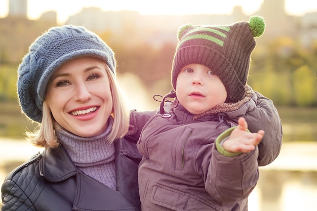 Mulher loira feliz e bonita de chapéu beijando filho bebê em uma jaqueta quente segurando-o em seus braços no parque outono