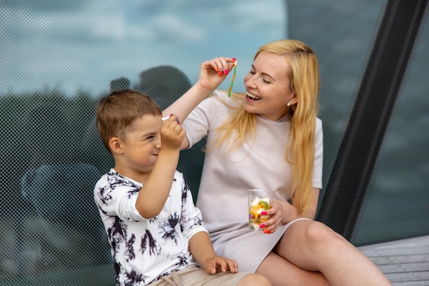 Mulher loira feliz e garotinho sentado no terraço e comendo doces Mãe e filho aproveitam o tempo juntos Mãe jovem positiva brincando passando tempo com seu filho fofo rindo se divertindo Família