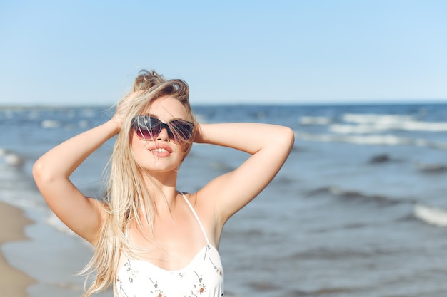 Mulher loira feliz em êxtase de felicidade livre na praia do oceano em pé com óculos de sol e posando