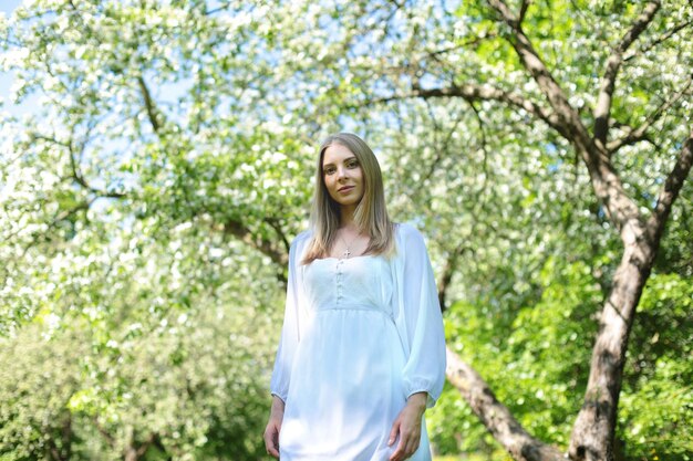 Mulher loira feliz em um vestido branco em um fundo de um jardim de primavera florescendo com as mãos levantadas para o céu