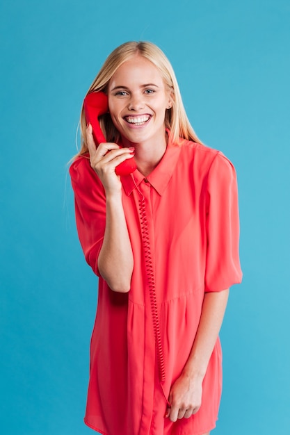 Mulher loira feliz falando no telefone vermelho e olhando para a frente, isolada em uma parede azul
