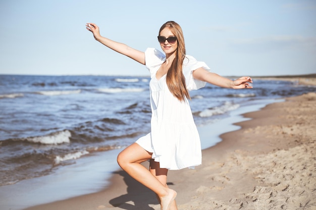 Mulher loira feliz se divertindo na praia do oceano em um vestido branco e óculos escuros.