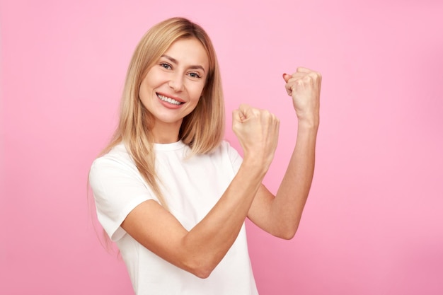 Mulher loira feliz sorrindo comemorando a vitória se alegra cerrando os punhos isolados no fundo rosa