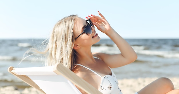 Mulher loira feliz usando óculos de sol e relaxando em uma cadeira de madeira na praia do oceano