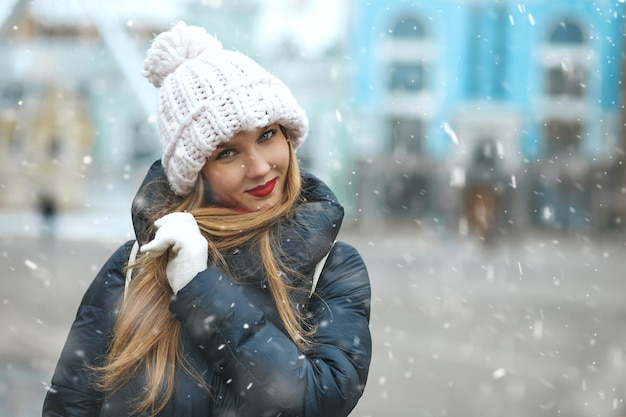 Mulher loira feliz vestindo boné de malha, caminhando na cidade durante a queda de neve. Espaço para texto